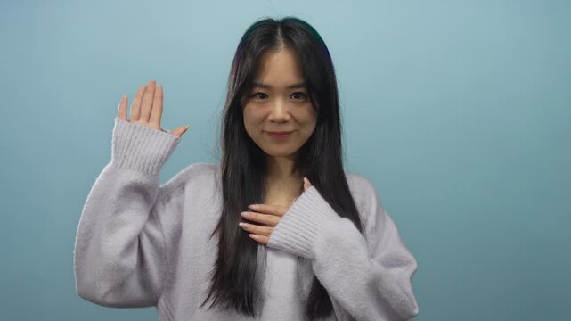 Young chinese woman makes oath gesture against isolated blue background, wearing cozy sweater, looking forward with confident expression, showcasing promise concept in a clear setting.