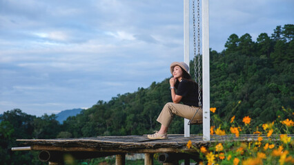 Portrait image of a woman sitting on a swing with a beautiful flower garden and mountain on cloudy day