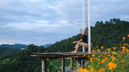Rear view image of a woman sitting on a swing with a beautiful flower garden and mountain on cloudy day