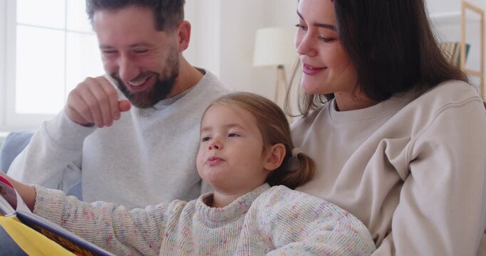 Happy cheerful parents having fun with cute child daughter reading book, album watching, playing together, mom and dad laughing embracing little kid daughter, funny enjoying family lifestyle at home