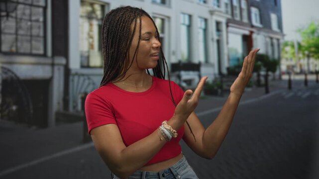 Woman gesturing with open hands on a street in front of row houses, wearing red crop top and jeans, braided hair visible; confidence openness.