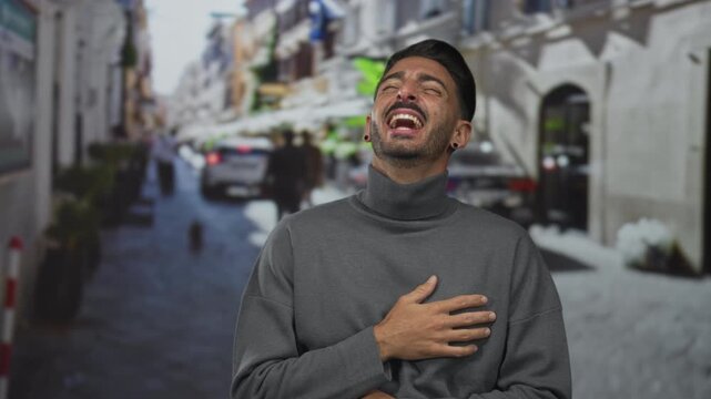 Man holds hand on chest and laughs broadly on narrow sunlit city street lined with blurred buildings; joy.