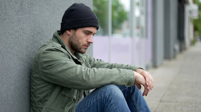 Lonely young man sitting on urban sidewalk against gray wall looking down in thoughtful depressed mood on an overcast day