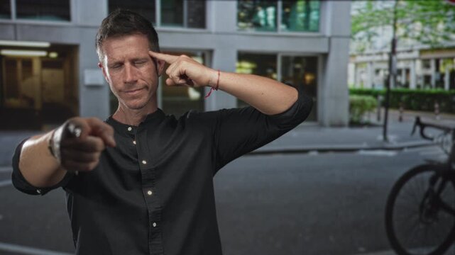 Man points finger to camera on urban street, finger touching temple in playful gesture; confidence challenge.