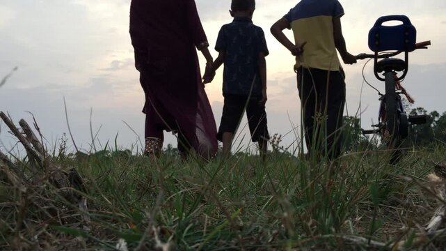 Atmospheric sunset footage showing children in silhouette with a bicycle against a warm evening sky. The sequence captures childhood, cycling, family bonding, outdoor recreation, freedom