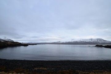 ヨーロッパの広大な湖と遠くに広がる雪山を望む曇り空の風景 北欧やアイスランドを思わせる静寂で神秘的な大自然のパノラマ 水面に映る空と山々のシルエットが印象的な旅行や観光のイメージ素材