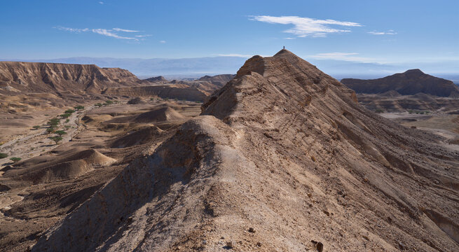 A sharp mountain ridge rises above wide and open valley of Nahal Zinin or Zanin in the Negev. A pair of tourists stands on a cliff with a panoramic view of the valley. Panoramic desert landscape.