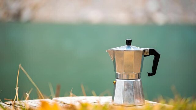 A metallic espresso maker sits on a wooden surface, with blurred natural greenery in the background