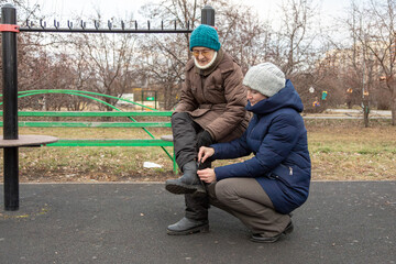 Woman helps an elderly woman buckle her boot while sitting on a park bench. Support and care for senior people in daily life. Intergenerational connection.
