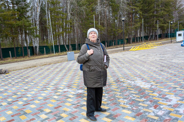 Elderly woman with book and backpack standing in autumn suburban park, looking around. Leisure activity for cognitive health.
