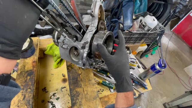 Close-up overhead POV shot of a mechanic wearing black gloves, holding scooter engine and turning the flywheel by hand to observe timing chain movement on a wooden bench in a home garage