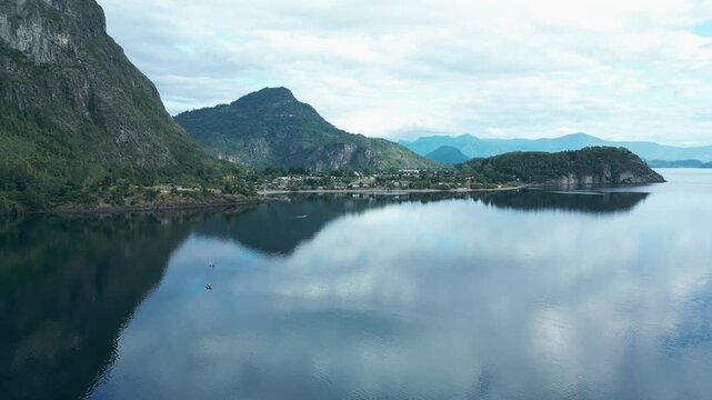 Forward drone aerial of calm lake with mirror reflections, dramatic rocky mountain cliffs backdrop, and serene wilderness peninsula