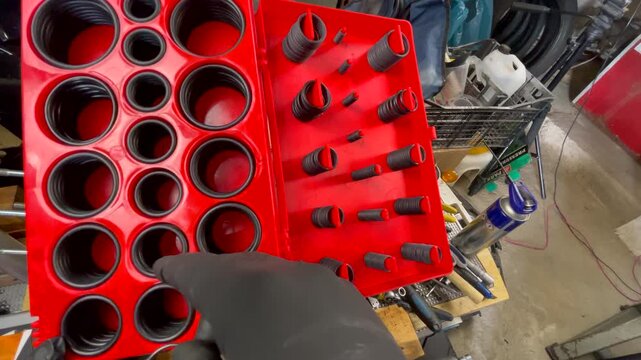 Close-up of mechanic wearing a black glove selecting the correct O-ring size from a red rubber O-ring kit, with tools visible on a home garage workbench