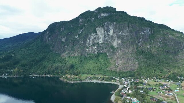 Backward drone aerial of dramatic rocky mountain cliff overlooking Ranco Lake shoreline village with houses and green forest landscape