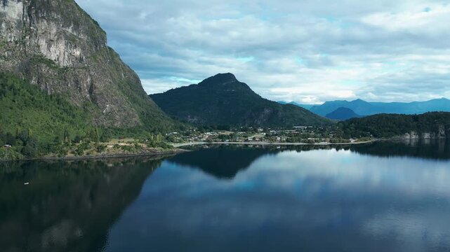 Orbiting drone aerial of Ranco Lake with mirror reflections, dramatic rocky cliffs, mountains backdrop, and Chilean village panorama