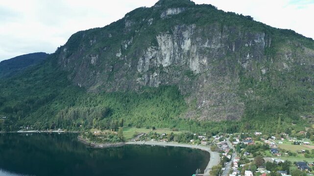 Revealing drone aerial of dramatic rocky mountain cliff towering over Ranco Lake shoreline village with houses and green forest