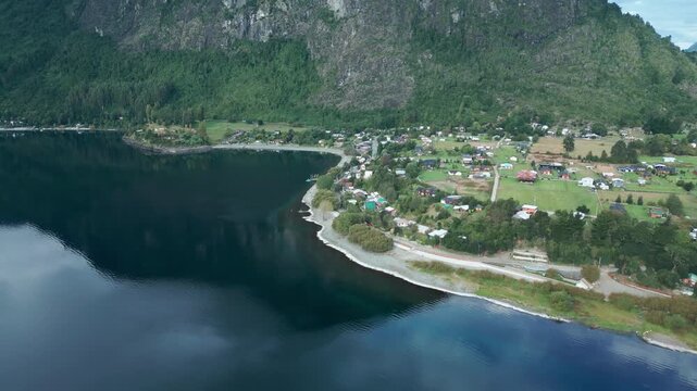 Forward drone aerial of Ranco Lake shoreline with coastal village, colorful houses, beach, rocky cliffs backdrop, and rural landscape