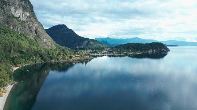 Forward drone aerial of Ranco Lake with mirror reflections, dramatic rocky cliffs, peninsula, beach, and Chilean mountains panorama
