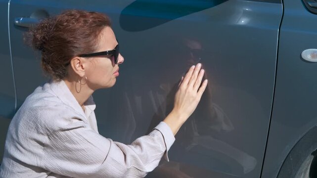 Woman examining scratches on car door. Woman wearing dark sunglasses carefully inspecting gray car exterior, scanning surface for potential scratches or dents under bright sunlight