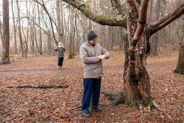 man looks at his watch while waiting for a woman who playfully sneaks up behind him in a park near a large tree. Concept of dating and relationships.