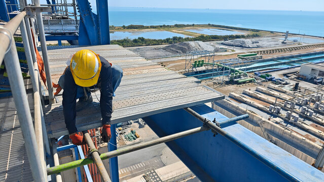 Construction worker wearing helmet and safety gear installing scaffolding at height on an industrial site near the sea, emphasizing teamwork, safety procedures, and offshore engineering environment.