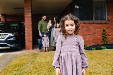 Close up of young girl standing outside house with family on the background