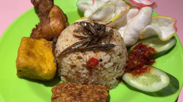 Traditional Indonesian rice dish with fried anchovies, tofu, tempeh, chicken, sambal chili paste, cucumber, and crackers on green plate