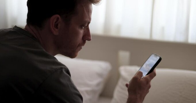 Man doom scrolling through social media or waiting for a message. Alone in bedroom, staring at smartphone. emotional distance psychological strain. Smartphone addiction. Digital overload, loneliness.