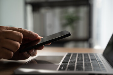 Hand operating a smartphone beside a laptop in an indoor workspace, using smartphone remote work and technology lifestyle with copy space background