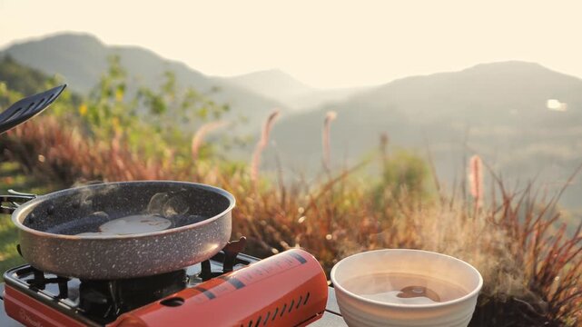 Outdoor cooking set on a mountain campsite at sunrise. This scenic image features a portable stove, pot, and bowl against a beautiful hazy valley, perfect for travel and adventure lifestyle themes.
