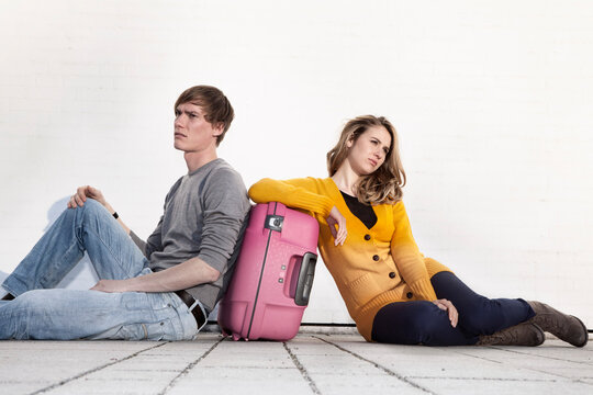 Couple waiting with suitcase sitting back to back outdoors