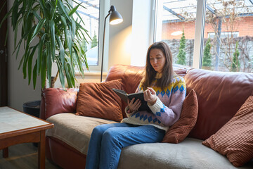 Fototapeta premium Woman reading a book while sitting on a comfortable sofa in a cozy living room