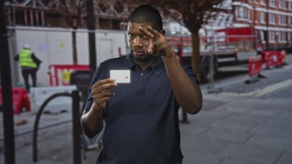 Man holds creditcard in one hand and makes a v finger gesture to his face while also pointing in other frames on a street; confidence trust.