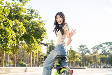 Happy asian woman waving hand while riding green rental bicycle on pavement in park or city square.