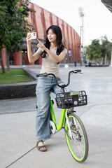 Pretty asian woman holding phone taking photos while riding on rental bicycle in park or city plaza.