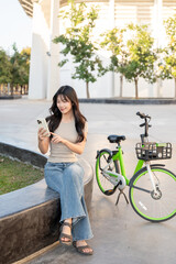 Pretty asian woman holding and looking at phone while sitting on bench aside bicycle in park plaza.