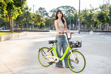 Laughing asian woman holding green rental bicycle standing on pavement in park plaza under sunlight.