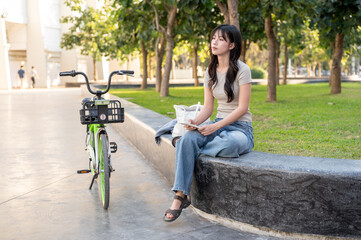 Pretty asian woman holding phone looking away as sitting on bench aside green rental bicycle in park