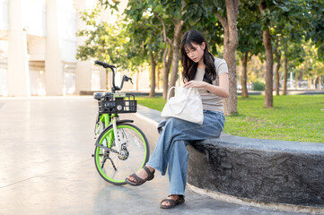 Pretty asian woman searching in her bag while sitting on bench aside a green rental bicycle in park.