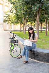 Pretty asian woman holding bag looking at phone while sitting on bench aside green bicycle in park.