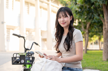 Pretty asian woman holding bag looking away from phone while sitting aside a green bicycle in park.