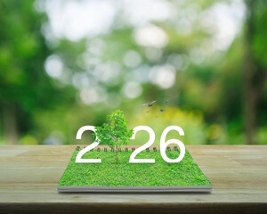 2026 white text with growing tree on green grass on open book on wooden table over blur green tree in park, Happy new year 2026 ecological cover concept