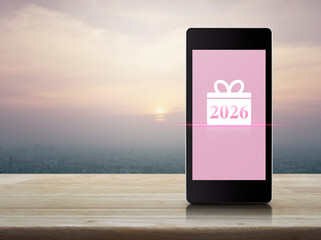 Gift box happy new year 2026 flat icon on modern smart mobile phone screen on wooden table over city tower and skyscraper at sunset, vintage style, Business happy new year 2026 shop online concept