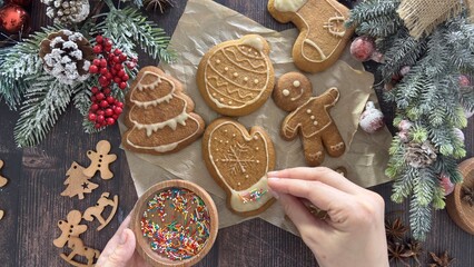 Fototapeta premium Gingerbread cookies of various shapes are laid out on the table. The maker decorates them with colorful sprinkles. Fir branches with pine cones and berries add a Christmassy feel. 