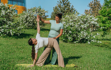 A yoga instructor guides a woman in a triangle pose in a sunny park, surrounded by flourishing blossoms and vibrant greens.