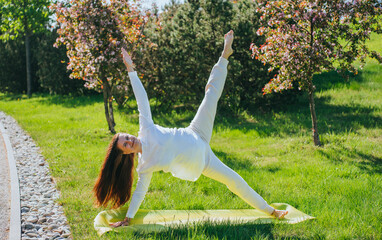 A woman balances gracefully on one hand and one foot in a yoga pose on a green mat, surrounded by blossoming trees and vibrant grass.