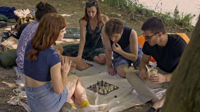 A cheerful group of young friends relax by the river, engaging in board games like chess and backgammon on a leisurely afternoon. They enjoy each other's company and the serene natural environment.