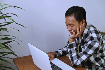 Young Gen Z man working remotely at home, thinking and focusing on tasks using laptop. Authentic daily life, modern lifestyle, and digital work concept.