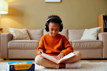 Black child sitting cross legged on floor wearing headphones reading book with focused expression, engaging in learning activity in living room with books nearby, looking down at pages