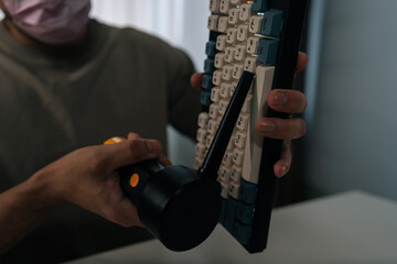 Cropped shot of man in protective mask cleaning mechanical wireless keyboard with electric air duster on desk, removing dust and debris for routine tech maintenance and hygienic workstation care.